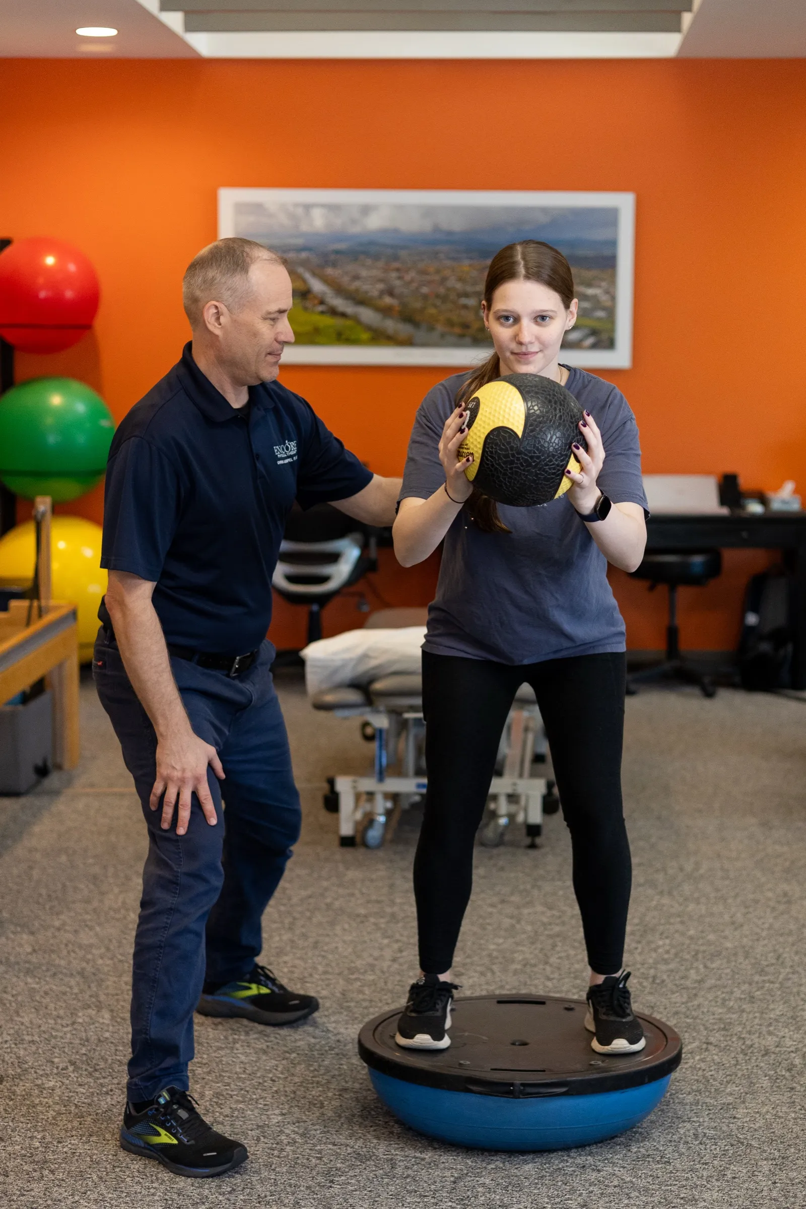 A patient training balance on a BOSU ball while holding a medicine ball, coached by an Encore therapist