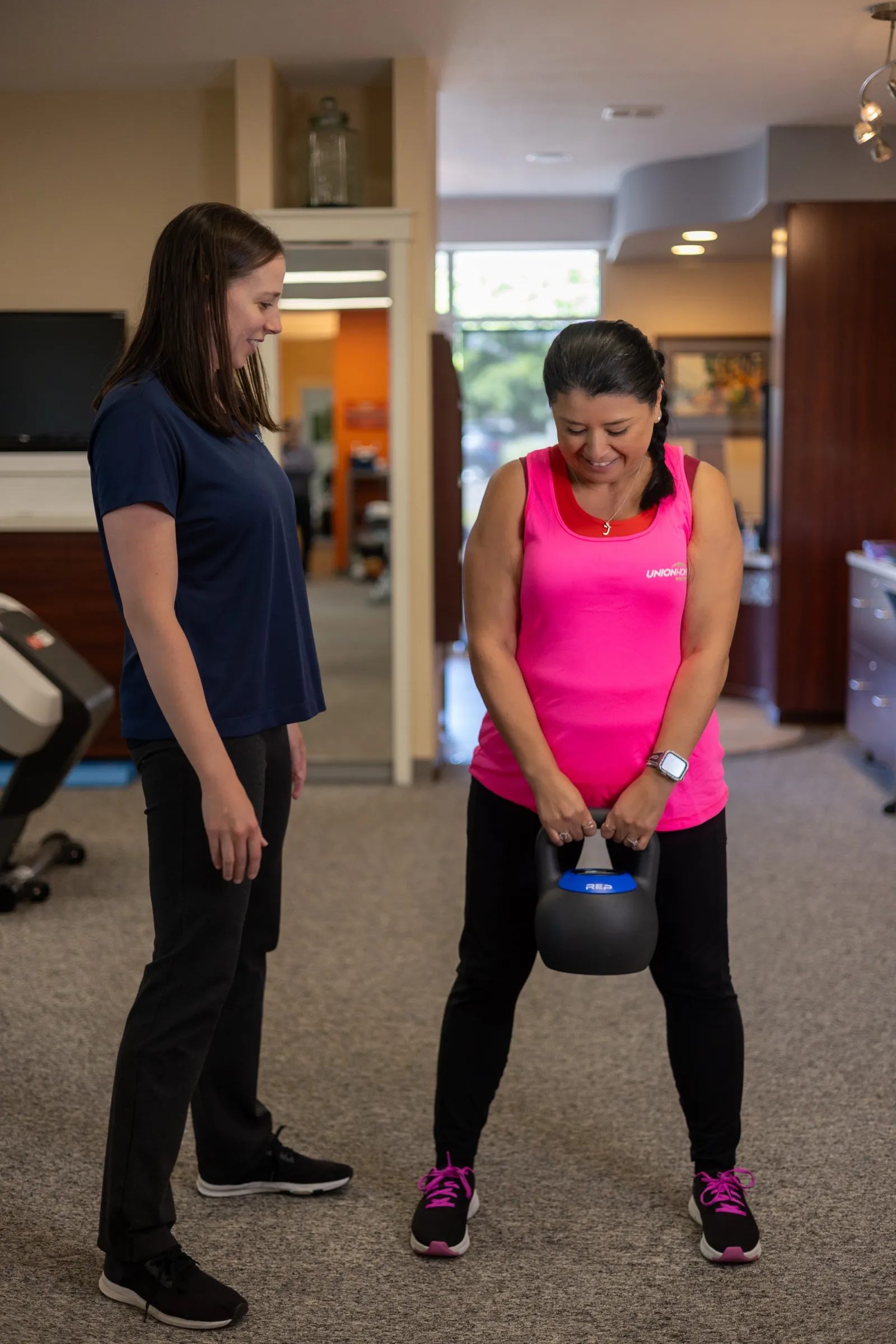 A smiling patient performing a kettlebell deadlift under an Encore therapist's supervision